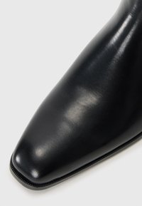 Bota de tobillo de piel negra con puntera afilada, textura suave y costuras decorativas a lo largo de la suela.