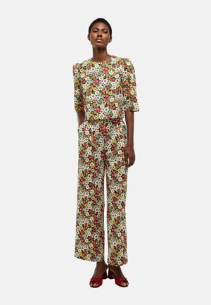 Femme debout portant un haut à imprimé floral assorti avec des manches au coude et un pantalon à jambes larges, associé à des sandales rouges à talons.