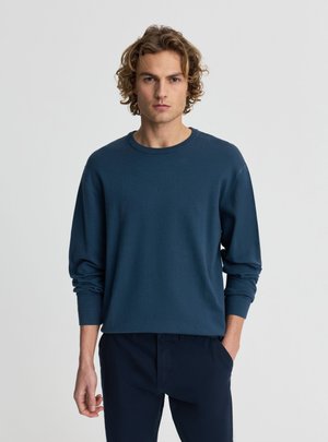 Giovane uomo con capelli biondi ricci che indossa un maglione blu scuro a maniche lunghe e pantaloni scuri su uno sfondo chiaro uniforme.