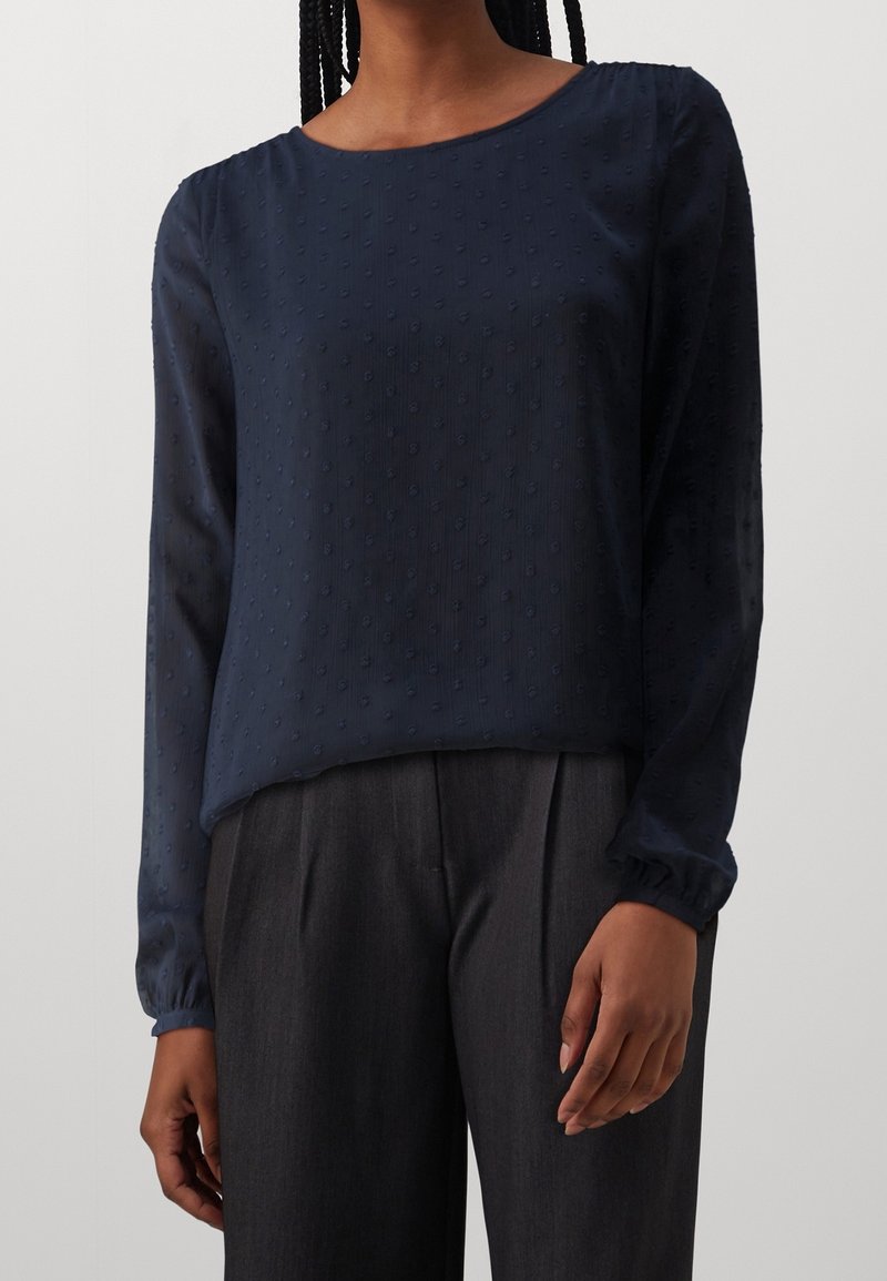 Femme portant un chemisier texturé bleu marine à manches longues avec un col rond, associé à un pantalon plissé gris foncé.