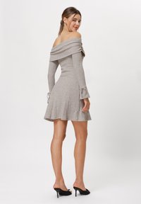 Off-shoulder geribd jurkje in lichtgrijs met lange mouwen en strikdetails bij de manchetten; uitlopende rok. Draagt het met zwarte hakken.
