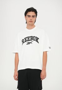 Bijela pamučna majica s velikim crnim logotipom "REEBOK" i malim logotipom na rukavu. Opušteni kroj s kratkim rukavima i okruglim izrezom.