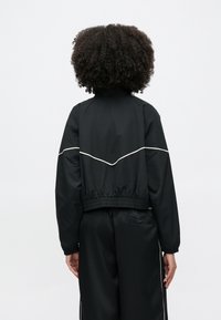 Μαύρο cropped μπουφάν με ελαστική μέση και μανσέτες· διαθέτει λευκή διακόσμηση κατά μήκος της πλάτης· λείο, ελαφρύ ύφασμα.