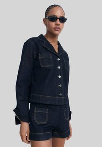 Veste en denim foncé avec un col cranté, fermeture à boutons et poches avant, accompagnée d'un short en denim assorti avec des poches arrière.