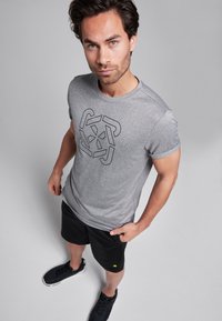 Grijze t-shirt met korte mouwen en een zwart geometrisch logo, gecombineerd met zwarte shorts en zwarte sportschoenen, tegen een neutrale achtergrond.