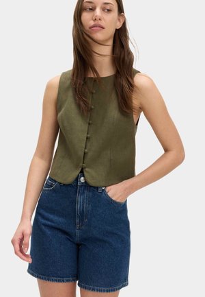 Femme portant un gilet sans manches vert olive boutonné et un short en jean bleu foncé taille haute, une main dans la poche, regardant sur le côté.