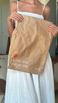 Persona con vestido blanco sosteniendo una bolsa arrugada marrón de Zalando con el texto "Ámame. Vísteme." visible en el frente.