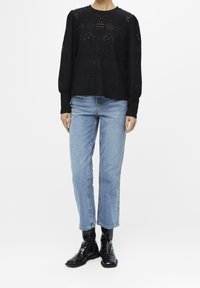 Blouse noire à manches longues texturée avec des motifs ajourés, associée à un jean droit bleu clair et des bottines noires brillantes.