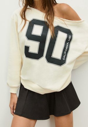 Frau trägt einen beigen Sweatshirt mit offener Schulter und großem schwarzem Aufdruck "90 California, USA" sowie einem schwarzen ausgestellten Minirock.