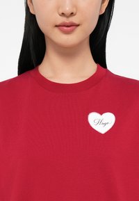 Femeie îmbrăcată cu un tricou roșu cu guler rotund, având pe piept un emblema în formă de inimă albă pe care este scris "Hugo" cu un font cursiv.