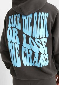 Sudadera negra de ajuste amplio, con gran texto azul que dice "TOMA EL RIESGO O PIERDE LA OPORTUNIDAD" en la parte de atrás. Textura suave.