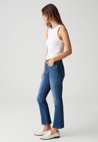 Top bianco senza maniche, jeans svasati in denim blu e mocassini bianchi. Il tessuto ha una texture liscia con minimi dettagli e cuciture a contrasto.