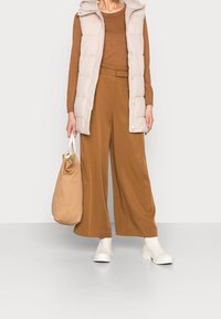 Beige padded vest over a brown long-sleeve top, paired with brown wide-leg trousers. White ankle boots and a beige bag complete the look.