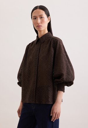 Kvinde med glat, mørkt hår iført en mørkebrun mønstret bluse med puffede ærmer og mørkeblå jeans, stående foran en ensfarvet baggrund.