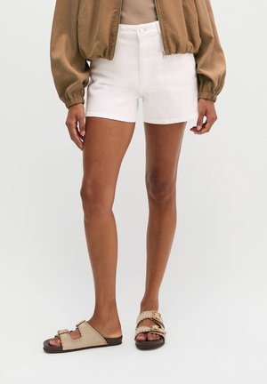 Person trägt weiße Shorts, beige Sandalen mit Schnallen und eine braune Jacke, steht vor einem einfarbigen hellen Hintergrund.