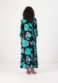 Robe maxi noire avec de grands motifs floraux turquoise et roses, à manches longues et à la coupe fluide. Portée avec des sandales à talons violettes.