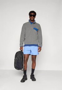Grauer Fleece-Pullover mit blauen Akzenten und Brusttasche, kombiniert mit hellblauen Shorts und schwarzem Rucksack. Schwarze Sneaker und Sonnenbrille getragen.