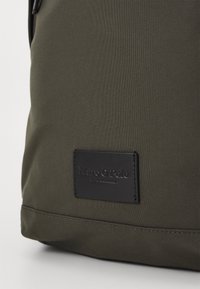 Zaino in tessuto verde con un patch in pelle nera con testo in rilievo, cuciture robuste e una superficie liscia.