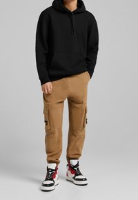 Černá mikina s kapucí, béžové cargo kalhoty s kapsami a černé, červené a bílé vysoké tenisky. Příležitostní streetwearová kombinace.