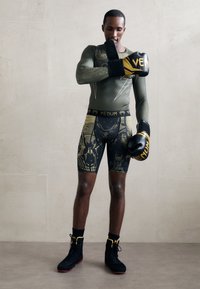 Schwarze und goldene Boxhandschuhe, olivgrünes langärmliges Oberteil, gemusterte schwarze Shorts. Schwarze Sportschuhe mit einem auffälligen Design und höherem Schaft.