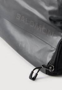 Κοντινό πλάνο γωνίας μαύρης τσάντας Salomon με λογότυπο της μάρκας και τράβηγμα φερμουάρ πάνω σε ανοιχτόχρωμη επιφάνεια.