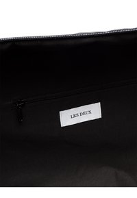 Juodas audinio maišas iš vidaus su užtrauktuku ir baltu etiketu, kuriame juodai atspaustas užrašas "LES DEUX". Satino tipo tekstūra ir minimalus dizainas.