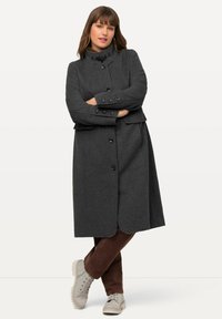 Cappotto grigio in misto lana con colletto alto, bottoni doppi e tasche laterali, abbinato a pantaloni in velluto a coste marroni e sneakers beige.