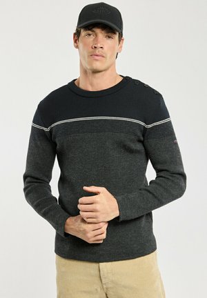 Mand iført en sort og mørkegrå sweater med skulderknapper og beige bukser, stående med hænderne foldet foran mod en ensfarvet baggrund.