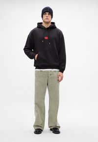 Felpa nera con cappuccio e logo rosso, pantaloni larghi verde chiaro, berretto nero con logo e scarpe nere. Stile casual, vestibilità rilassata.