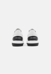 Baskets blanches avec un accent de talon noir portant la marque "STEVE MADDEN", dessus en cuir lisse et semelle en caoutchouc noir texturé.