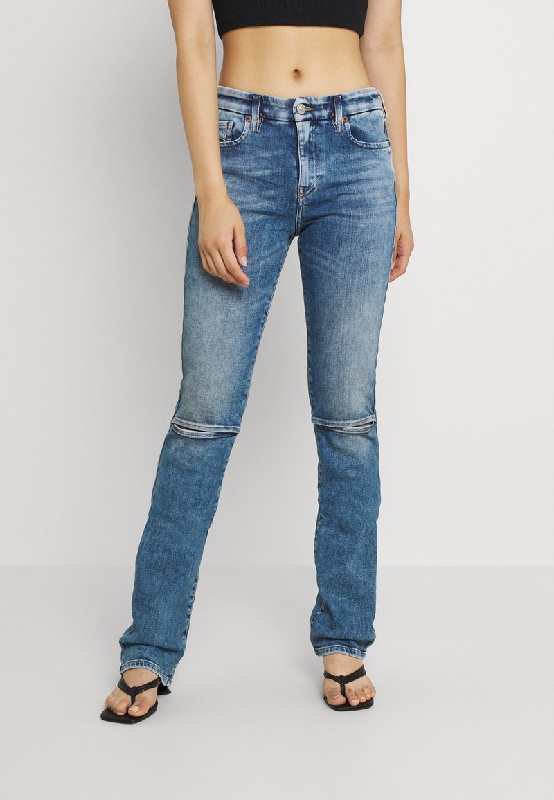 Femme portant un jean bleu lavage moyen avec des fentes aux genoux et des sandales noires à talons ouverts, debout devant un fond uni.