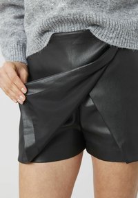 Shorts en cuir noir avec un design portefeuille, présentant une texture lisse et des accents brillants. La taille est cachée sous un pull gris clair.