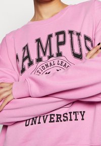 Sudadera rosa con texto negro que dice "CAMPUS" y "UNIVERSIDAD". Cuenta con puños y cuello acanalados, y está hecha de una tela suave y texturizada.