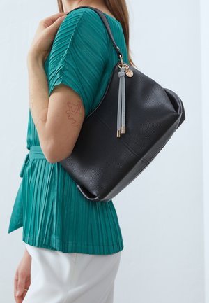 Femme en haut plissé vert sarcelle et pantalon blanc portant un sac à main en cuir noir texturé avec des détails à pompons gris sur l'épaule.
