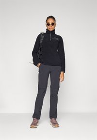 Pullover in fleece con mezza cerniera, nero; abbinato a pantaloni cargo grigi. Indossato con occhiali da sole e scarponi da trekking, completo di uno zaino.