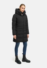 Long black puffer coat with a hood, featuring a zip closure, quilted design, and a knee-length cut. Paired with blue jeans and black boots.