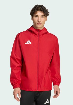 Joven con chaqueta roja con capucha y cremallera, y pantalones negros con logo blanco, de pie frente a un fondo claro.