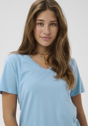 Mladá žena s dlhými hnedými vlasmi, oblečená v svetlomodrom tričku s výstrihom do V a vrstvami náhrdelníkov, jemne sa usmieva na jednoduchom pozadí.