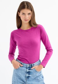 Top ajustado de manga larga en un tejido de color rosa brillante, combinado con unos vaqueros de cintura alta de color azul claro, manos en los bolsillos, fondo liso.