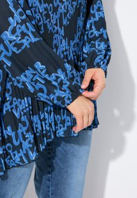 Top nero plissettato con motivi di testo astratti blu, maniche elasticizzate e vestibilità rilassata, abbinato a jeans in denim.