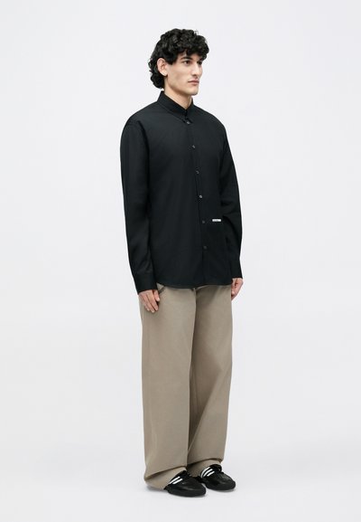 Chemise noire à manches longues avec fermeture à boutons, tissu léger, coupe droite, associée à un pantalon beige ample et des baskets noires.
