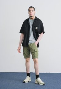Cămașă neagră cu mânecă scurtă peste un tricou gri, pantaloni scurți cargo de culoare verde măsliniu și adidași deschiși, stând pe un fundal alb simplu.