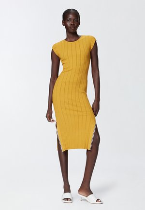 Abito midi giallo lavorato a maglia con texture a coste, maniche a balconcino, spacchi laterali e bordi decorativi. Abbinato a sandali bianchi con slide.