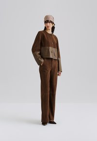 Giacca cropped in suede marrone con dettagli in pelle, abbinata a pantaloni marroni coordinati. Presenta grandi bottoni e una vestibilità moderna.