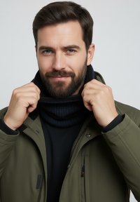 Dark green jacket with a high collar, black sweater underneath, and a textured dark scarf. The individual is adjusting the scarf around their neck.