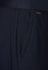 Marina ribbade byxor med en slät midja, sidoficka och en liten metalltagg. Tyget har ett vertikalt texturmönster.