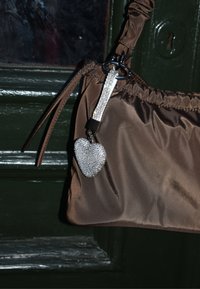 Geantă din țesătură maro cu design adunat, având un charm în formă de inimă cu cristale strălucitoare argintii și o curea texturată cu accente argintii.