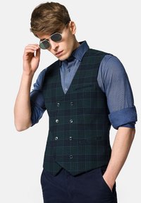 Zielona kamizelka w kratę z podwójnym zapięciem, srebrnymi guzikami i niebieską koszulą, noszona z ciemnymi spodniami. Srebrne okrągłe okulary przeciwsłoneczne.