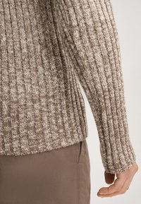 Brauner, gerippter Strickpullover mit einer gemischten Textur, der ein helles und dunkles Muster aufweist, kombiniert mit beigen Hosen. Fokus auf die Ärmeldetails.