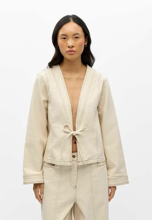 Femme aux longs cheveux foncés portant une veste beige nouée à la taille et un pantalon taille haute assorti sur un fond blanc uni.